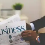 A person reading a business newspaper with financial charts visible.